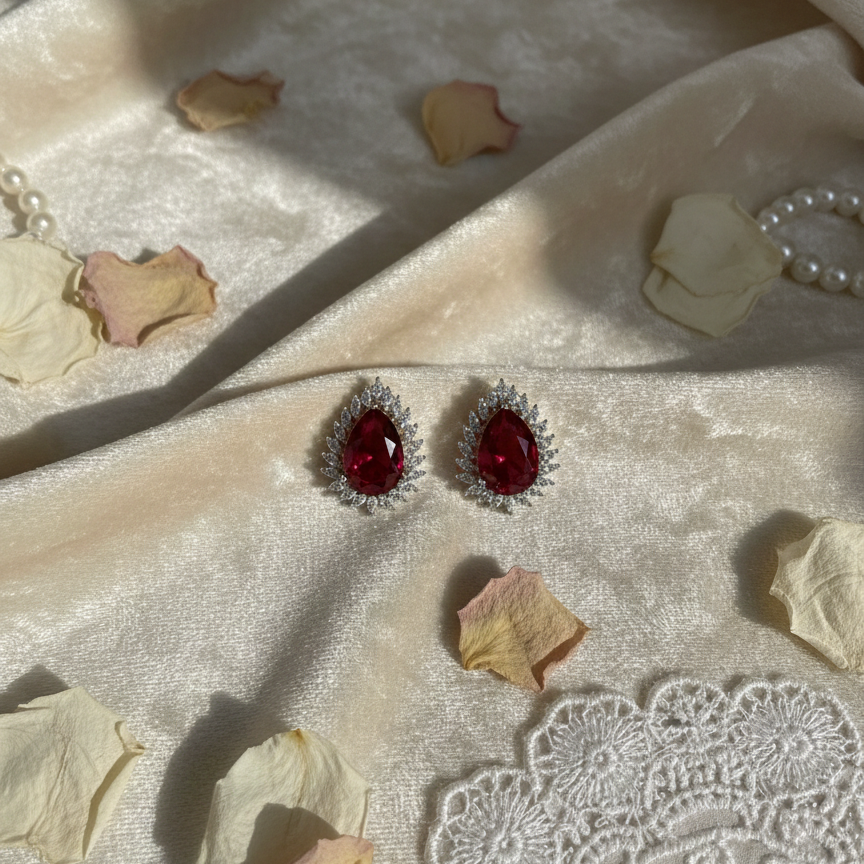 Red gemstone earrings on a textured fabric surface with dried flowers and lace.